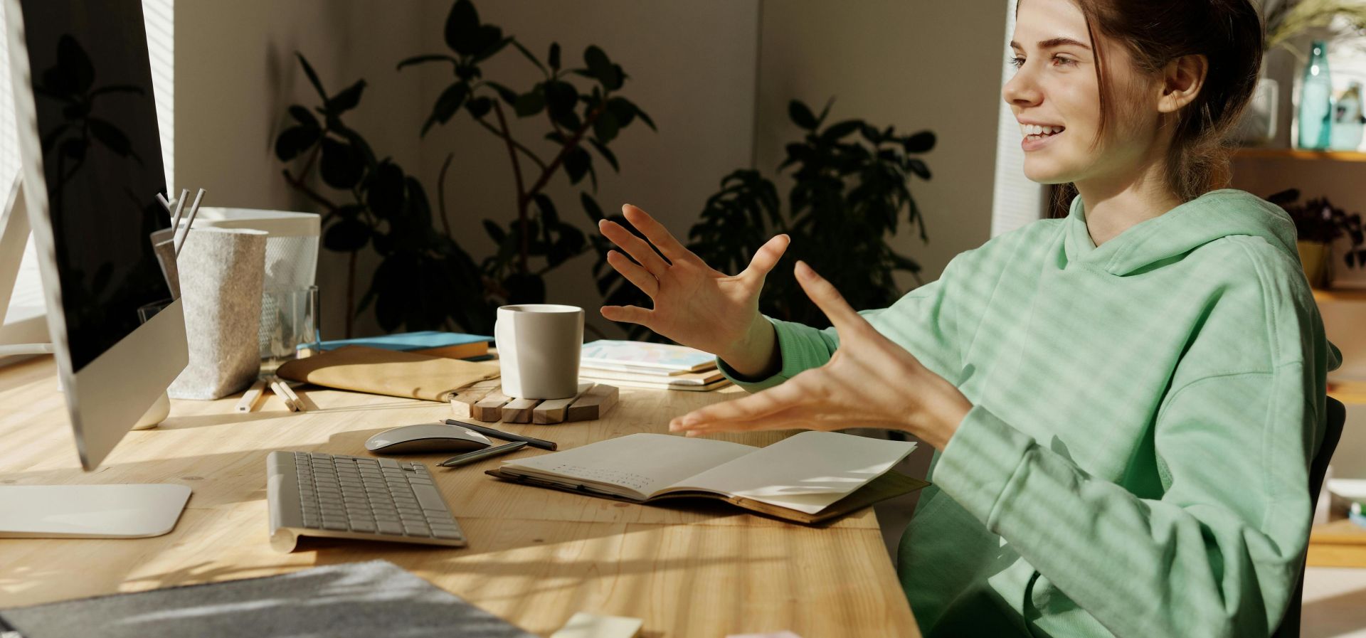 A woman in a green hoodie having a video call at her home office desk.