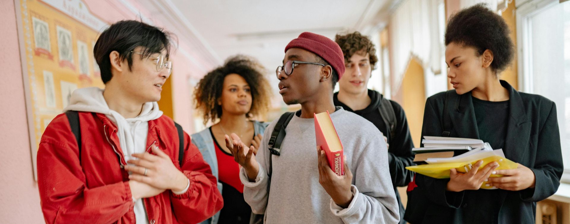 A group of diverse students walking and talking in a school corridor, carrying books and folders.