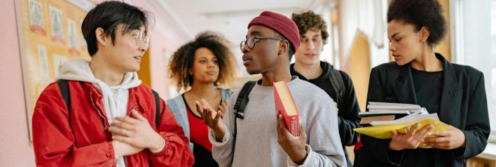 A group of diverse students walking and talking in a school corridor, carrying books and folders.